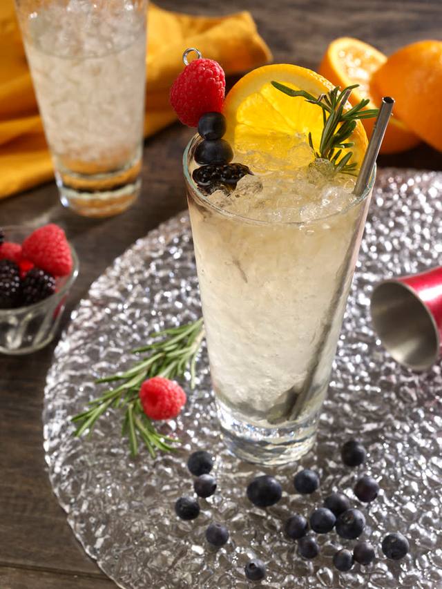 Collins glass of a sparkling cocktail over crushed ice garnished with berries, an orange wheel and rosemary
