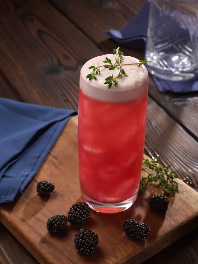 Tall glass of blackberry thyme fizz with a foam cap and thyme sprig on a wooden board