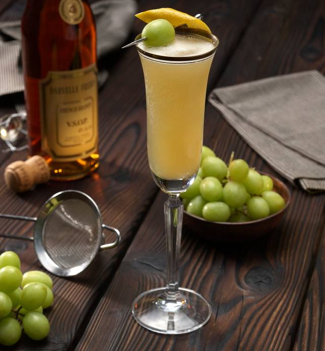 Tall flute of a brandy cocktail garnished with a green grape and lemon twist