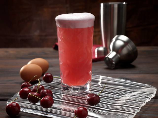 Tall glass of pink cherry fizz with a thick foam cap beside fresh cherries