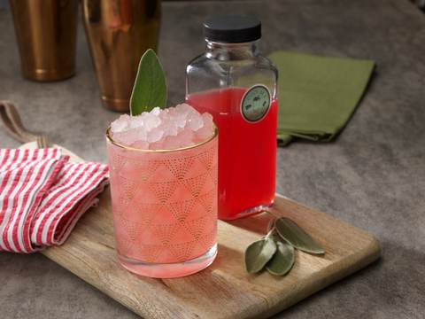 Cherry and sage cooler over crushed ice beside a bottle of house made syrup
