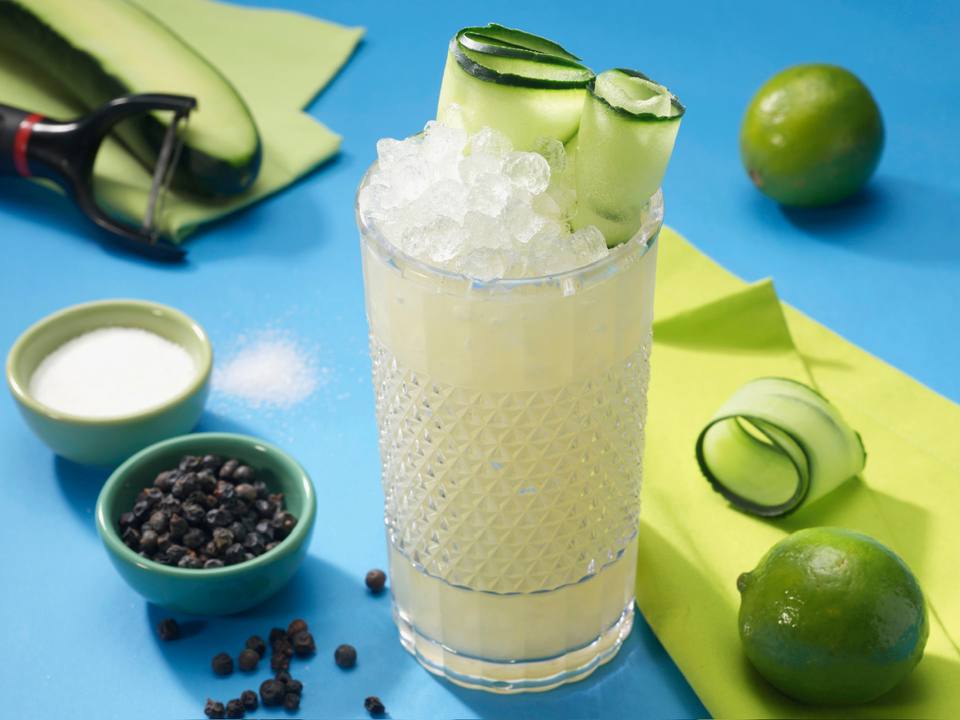 Textured highball of a cucumber gimlet over crushed ice with cucumber ribbons and limes