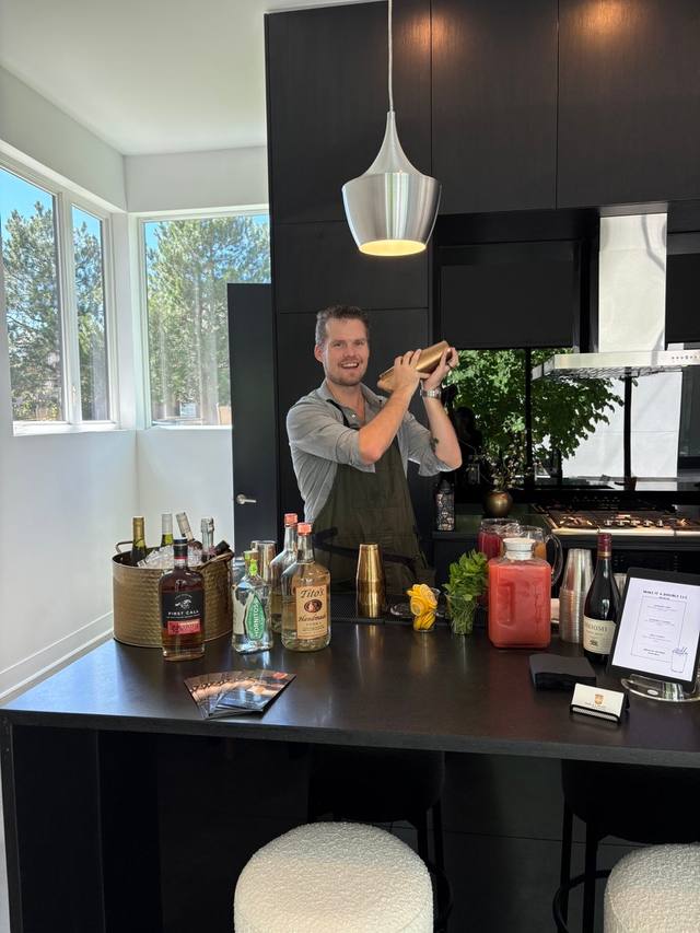 Bartender shaking a cocktail behind a home kitchen island bar set with spirits and glassware