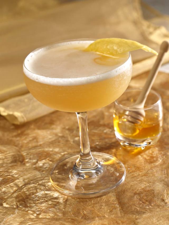 Coupe of a honey lemon sour with a foam cap and lemon twist beside a jar of honey