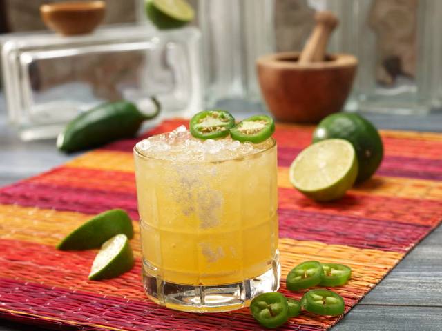 Rocks glass margarita over crushed ice topped with jalapeno slices beside fresh limes