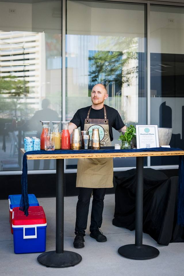 Bartender standing behind an outdoor mocktail station with juice carafes and a printed menu