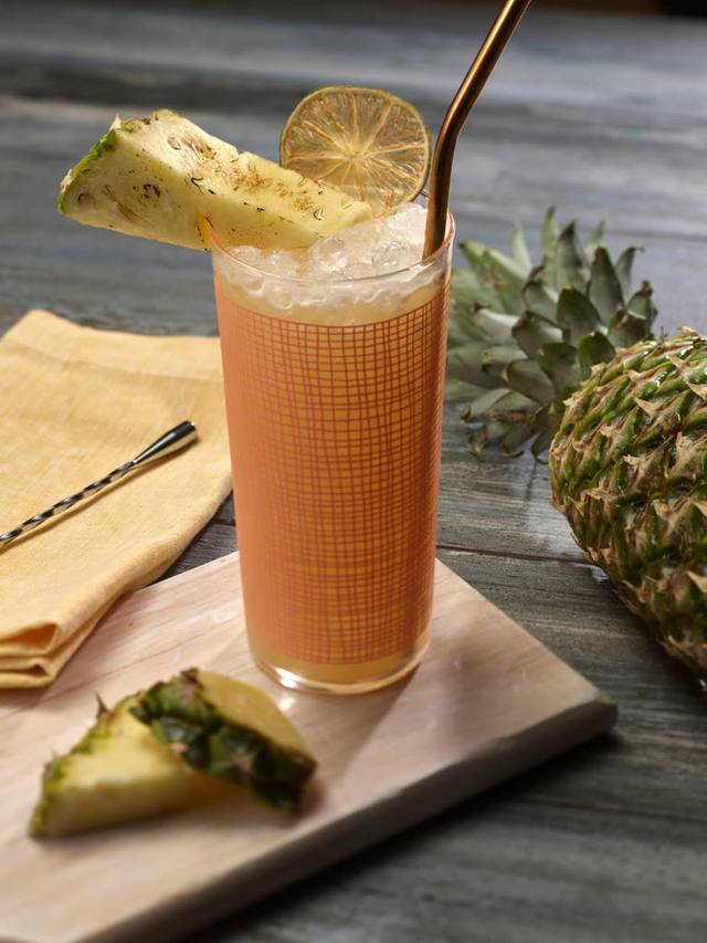 Tall patterned highball of pineapple cocktail over crushed ice with a charred pineapple wedge and lime wheel