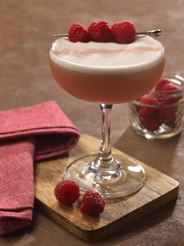 Coupe of raspberry sour with a silky foam top and three raspberries on a pick