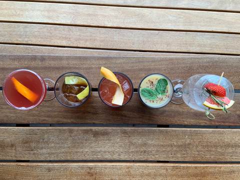 Overhead row of five garnished cocktails lined up on a wooden rooftop table