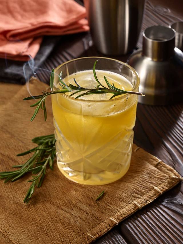 Cut crystal rocks glass of a golden sour with a smoking rosemary sprig