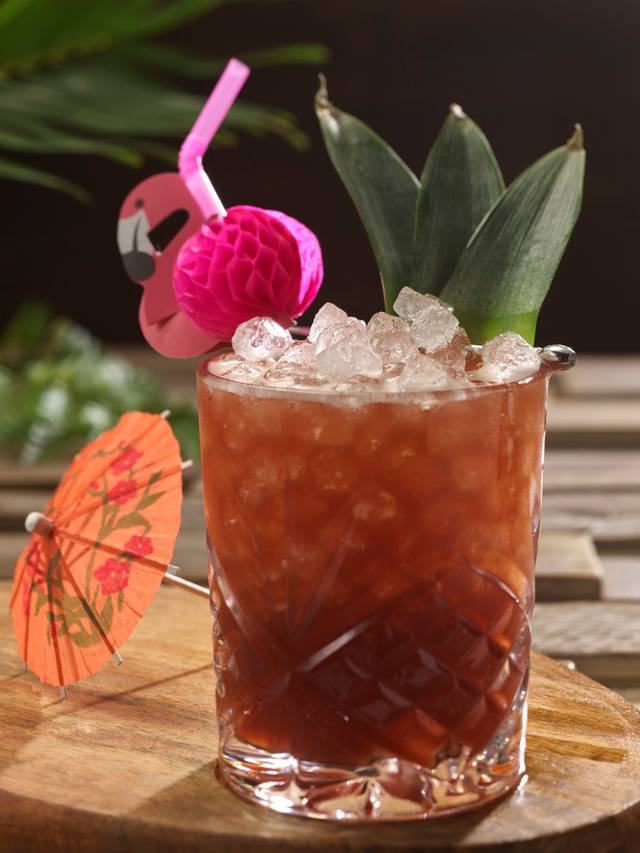 Cut glass tumbler of tiki rum punch over crushed ice with tropical leaves and a flamingo pick